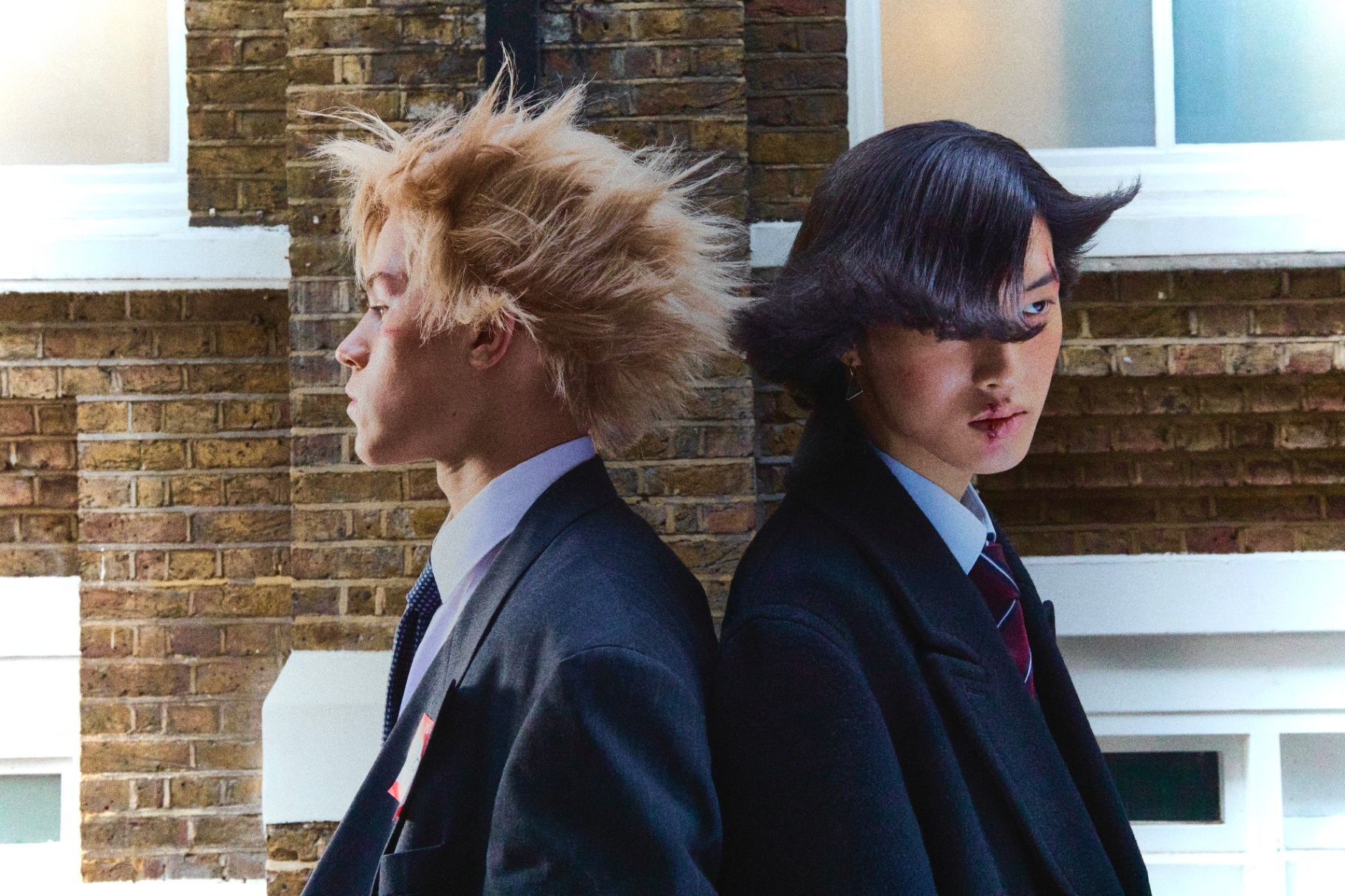 Two suited figures with styled hair against a brick wall